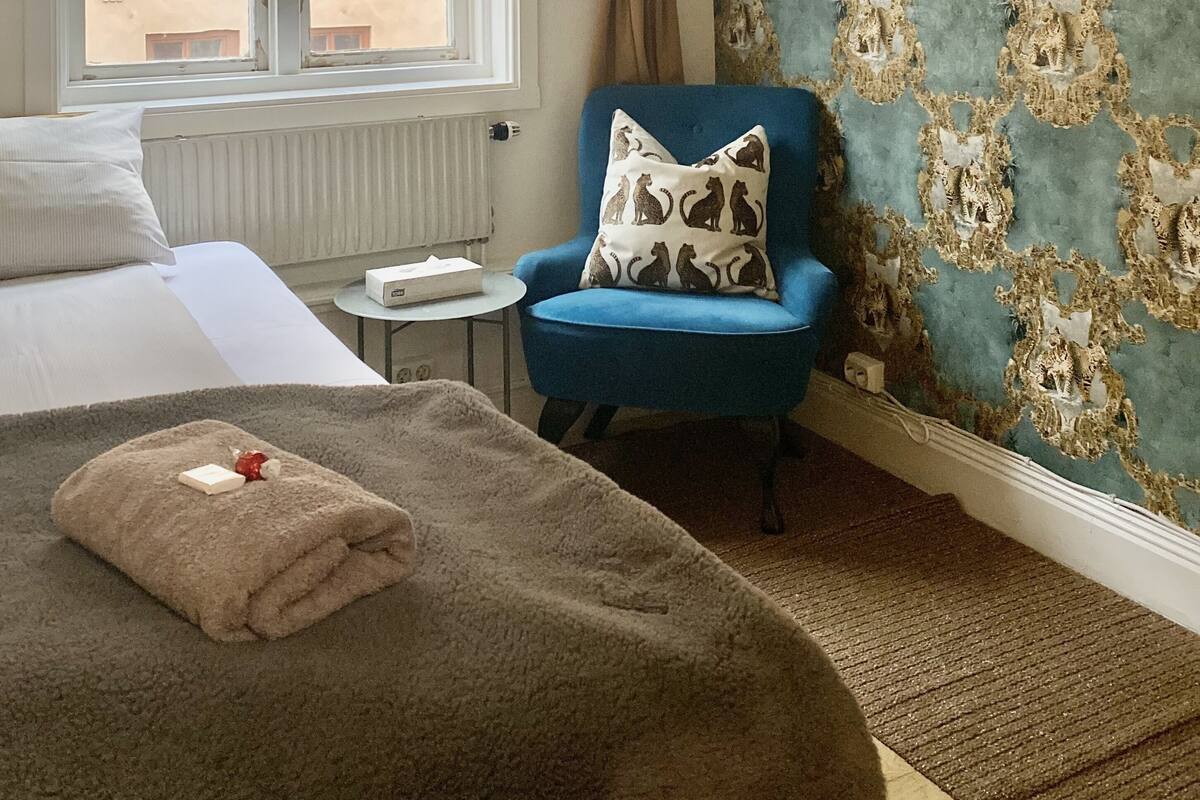 Una habitación de hotel con una cama, un sillón azul, un papel tapiz estampado y una ventana con cortinas.