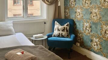Una habitación de hotel con una cama, un sillón azul, un papel tapiz estampado y una ventana con cortinas.