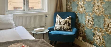 A hotel room with a bed, a blue armchair, a patterned wall, and a window with curtains.