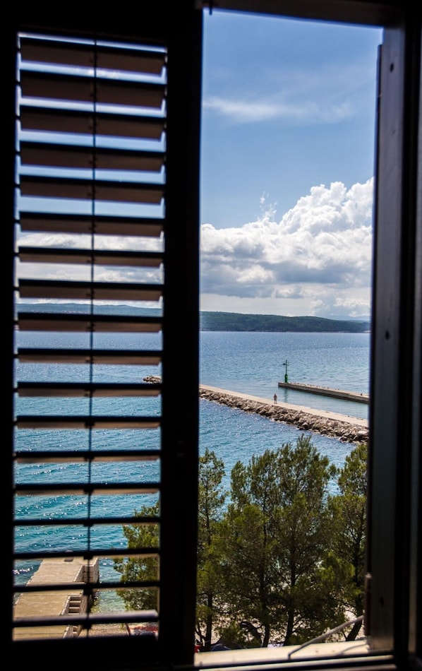 Paesaggio costiero visto attraverso una finestra con persiane orizzontali, un molo roccioso che si protende verso il mare e una costa lontana con alberi.