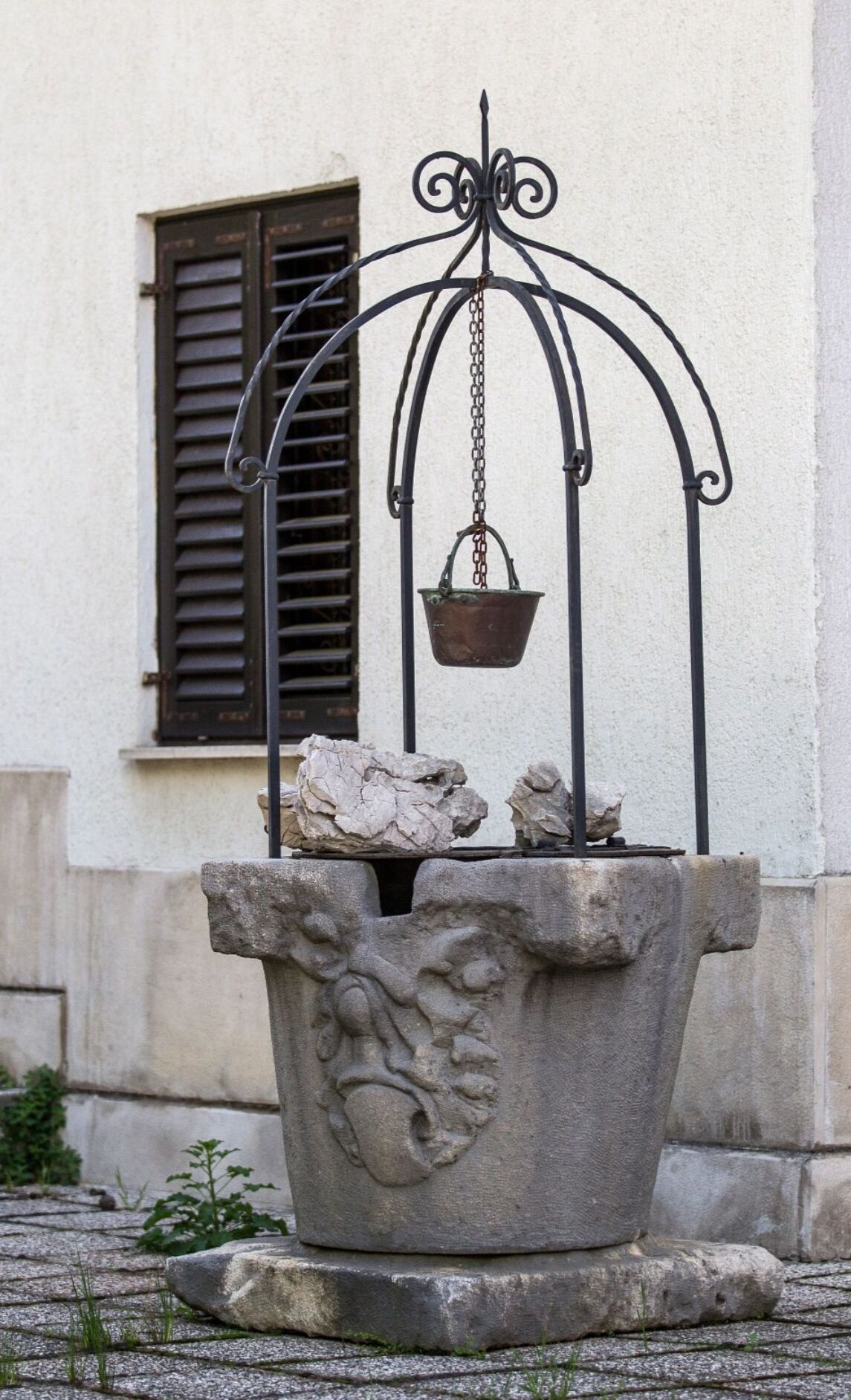 A stone fountain with a metal structure and a hanging bucket, set against a building with a window and shutters.
