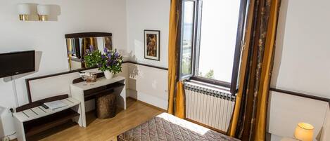 A hotel room with a bed, a desk with a chair, a TV, a window with curtains, and a radiator.