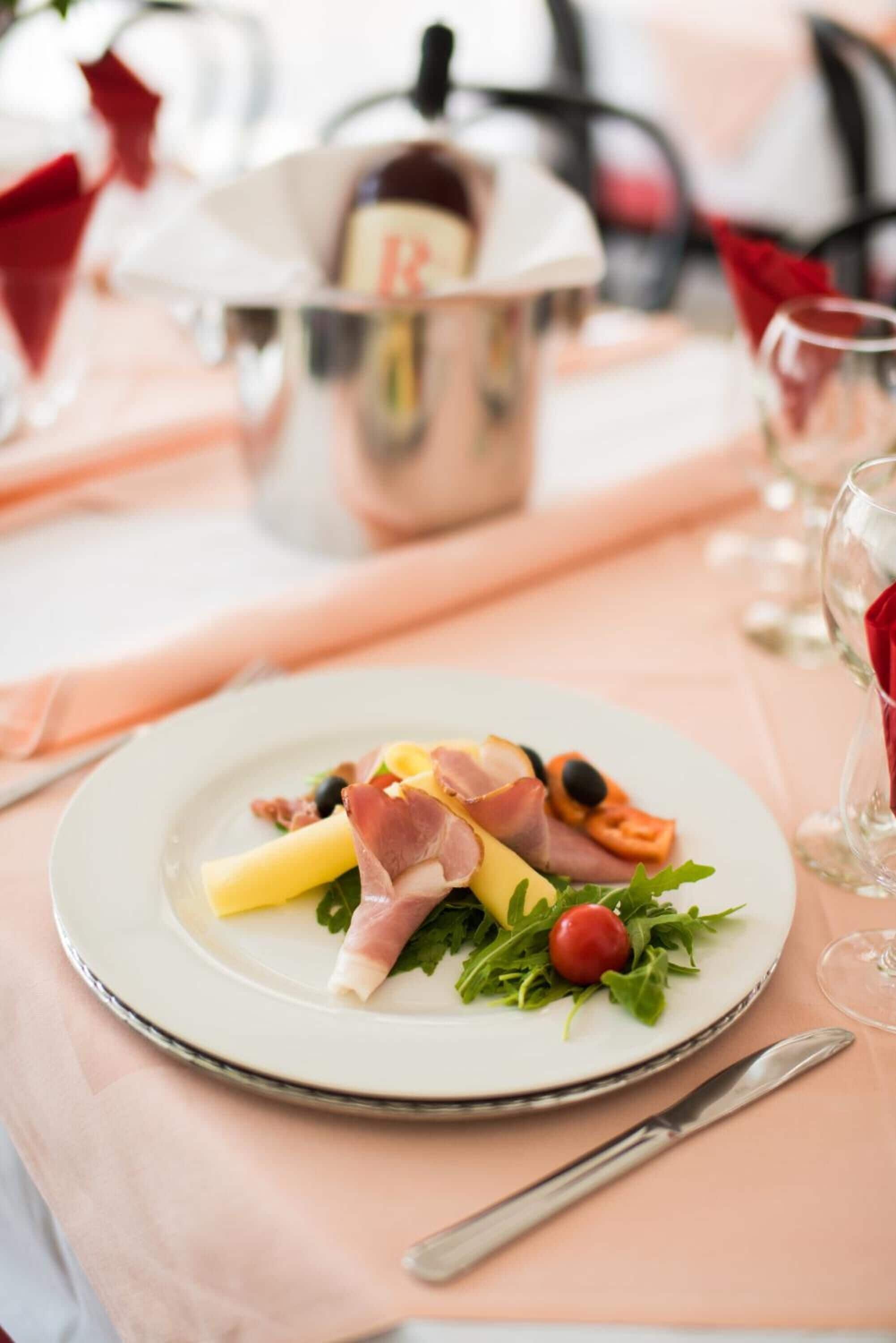 A plate of food with ham, cheese, and greens on a table with a bottle of red wine and glasses.