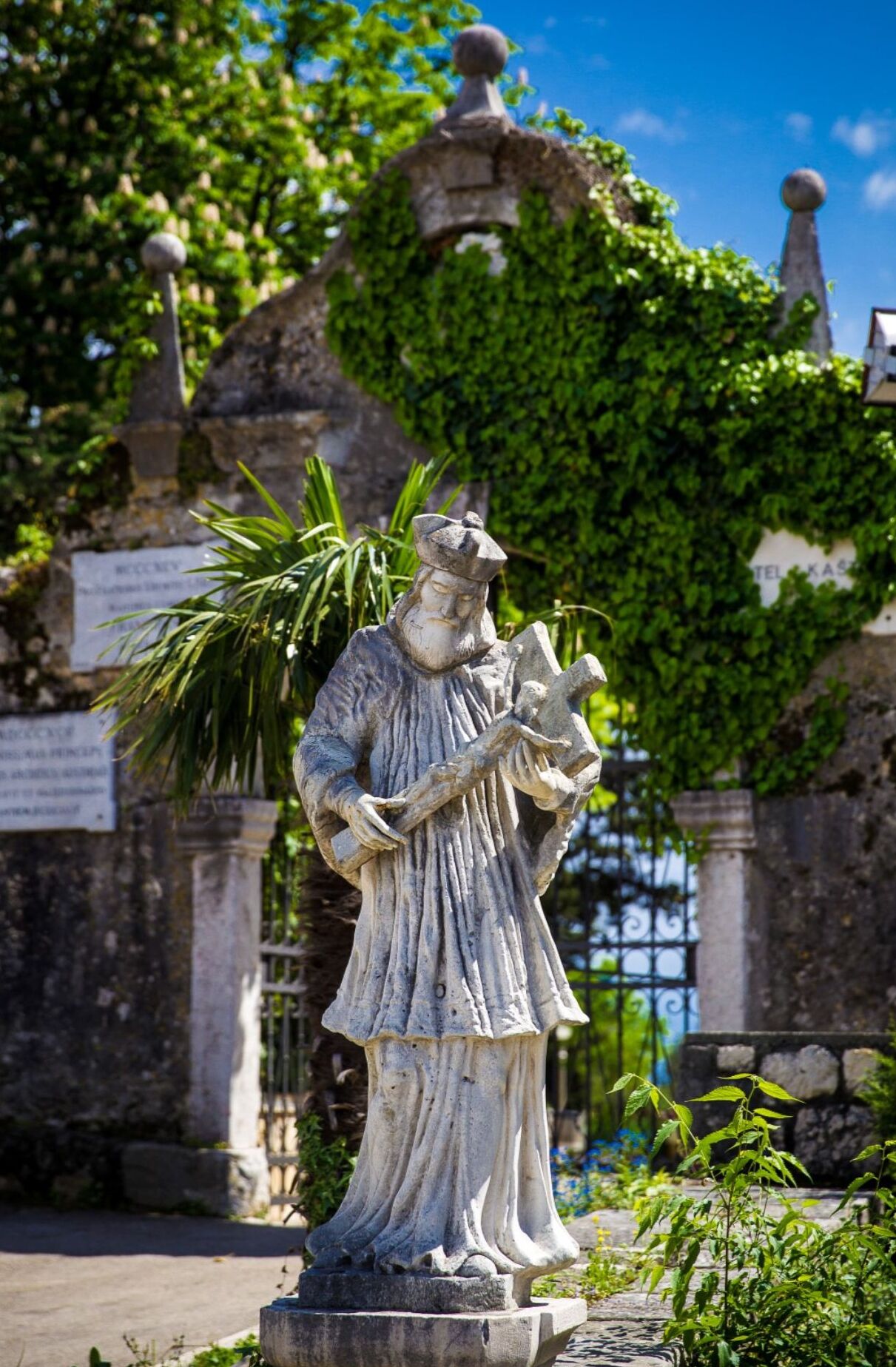 A statue of a figure holding a book, standing in front of a stone structure with a gate and greenery.