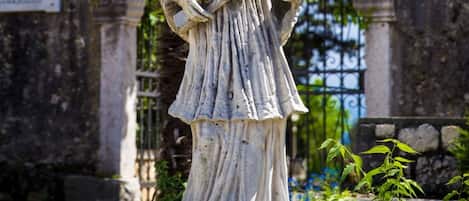 A statue of a figure holding a book, standing in front of a stone structure with a gate and greenery.