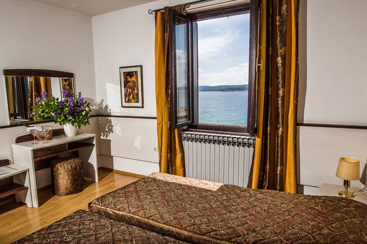 Hotellihuone, jossa on sänky, työpöytä, tuoli, ruukkukasvi, ikkuna verhoilla ja näkymä merelle.