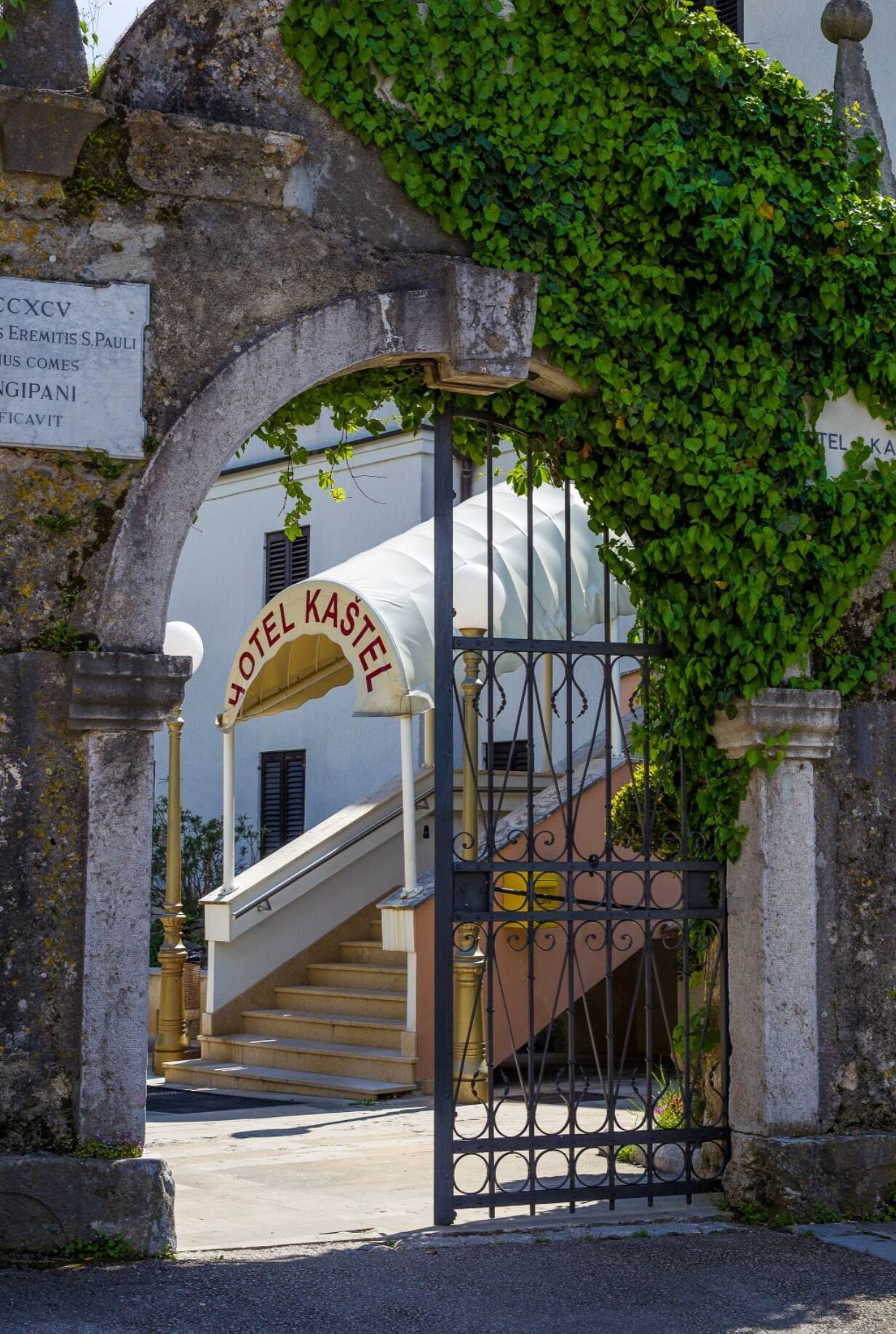 An entrance to Hotel Kastel with a stone archway, a staircase leading to the entrance, and a sign indicating the hotel's name.