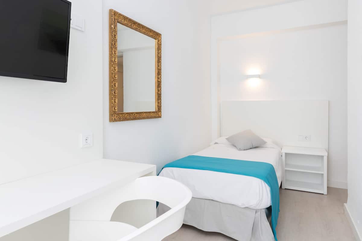 A modern hotel room with a single bed, a bedside table, a desk with a chair, a large mirror, and a flat-screen TV mounted on the wall.