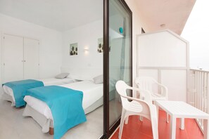 A room with two beds, a balcony with a table and chairs, and a glass door leading outside.