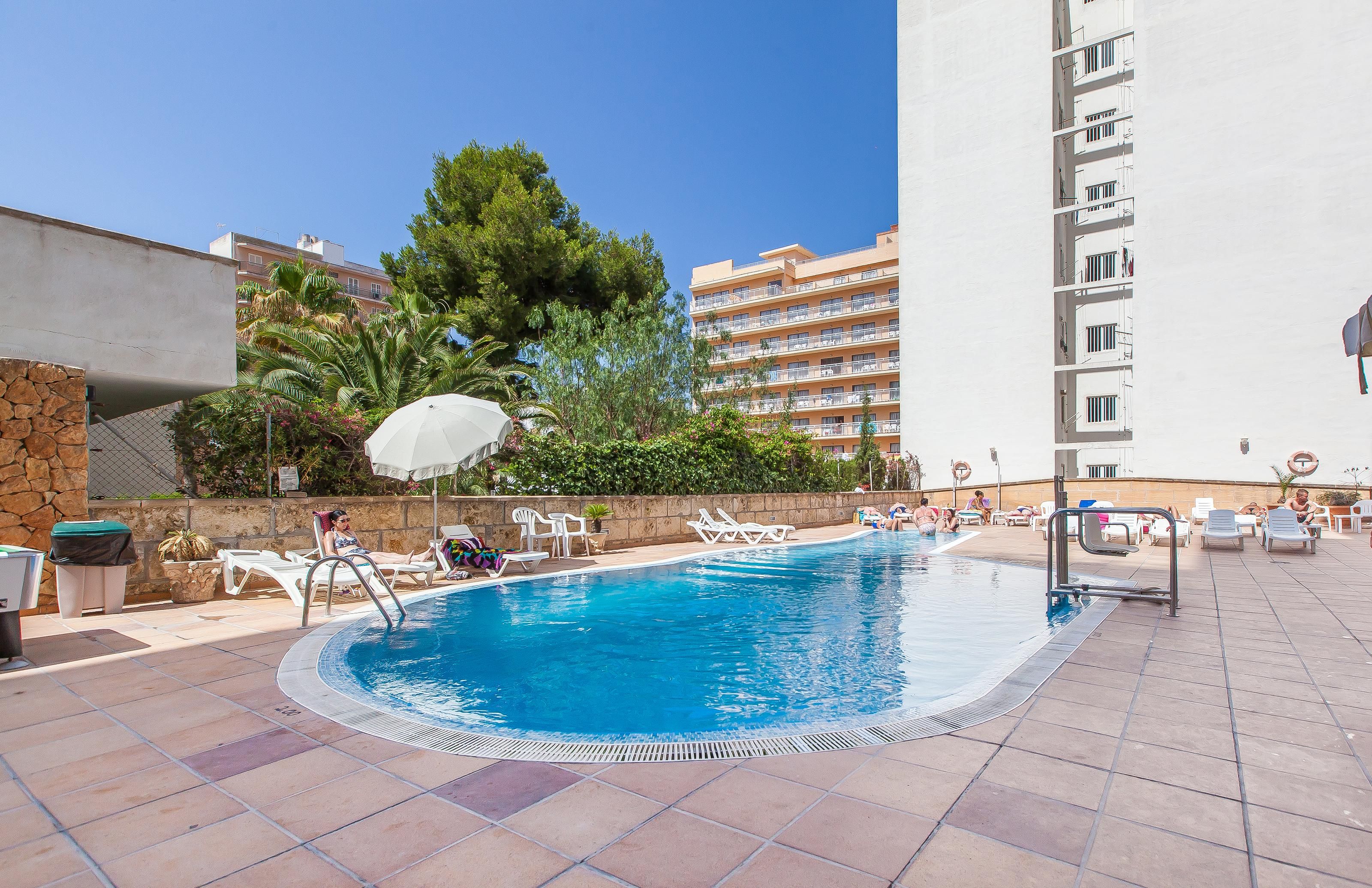 Una piscina con sillas de descanso y sombrillas, rodeada por un muro de piedra y follaje.