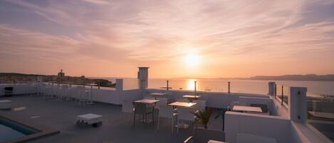An outdoor terrace with tables and chairs, overlooking a sunset over the sea.