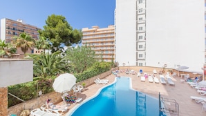 A pool area with lounge chairs and umbrellas, surrounded by greenery and a tall building. - BLUESEA Arenal Tower (Llucmajor)