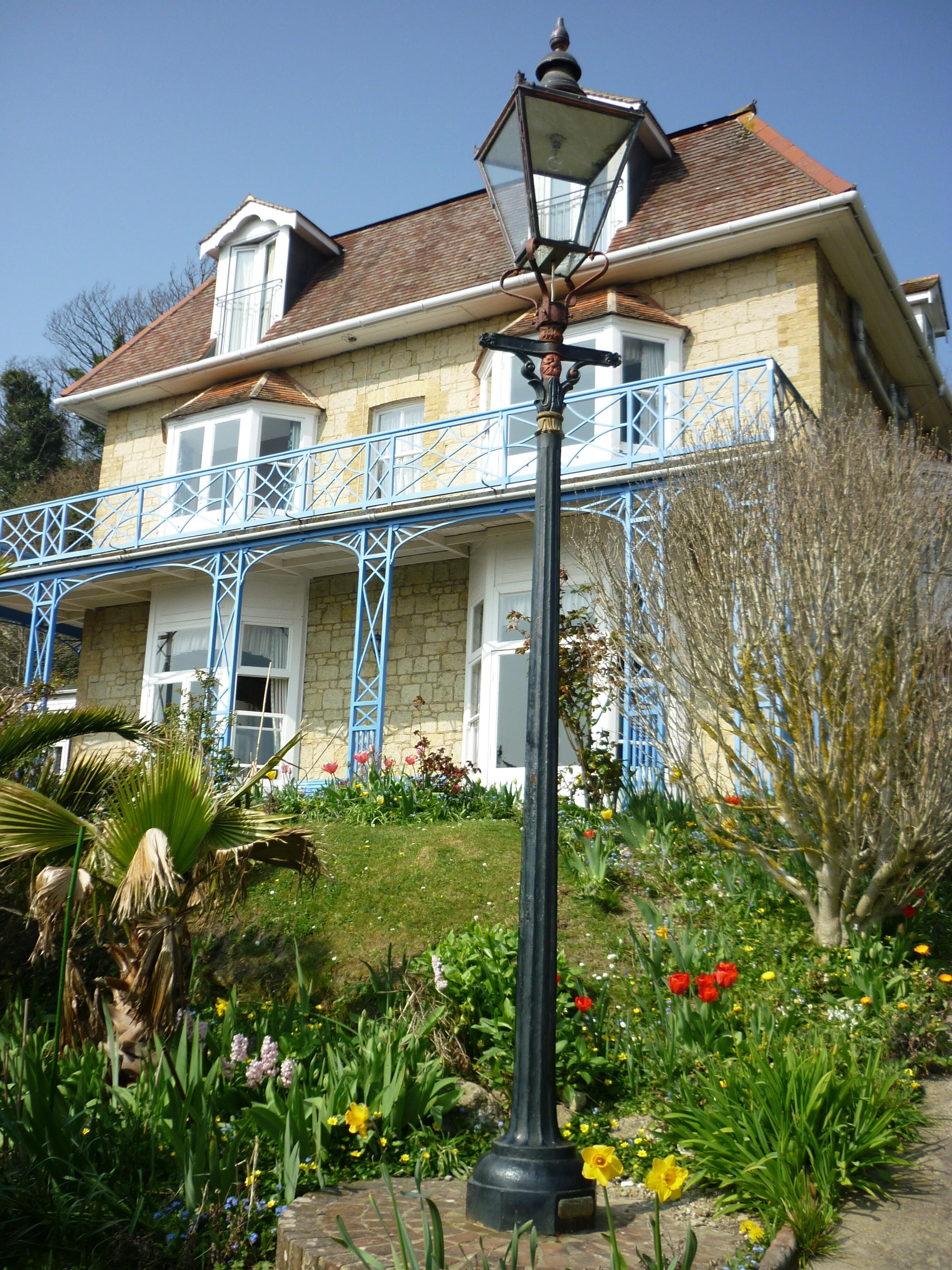Una casa de dos pisos con balcón, rodeada por un jardín con diversas flores y plantas.
