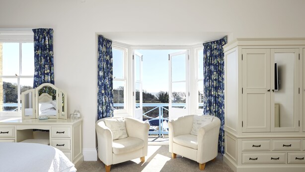 A bedroom with a bed, two white armchairs, a white dresser, and a large window with curtains.