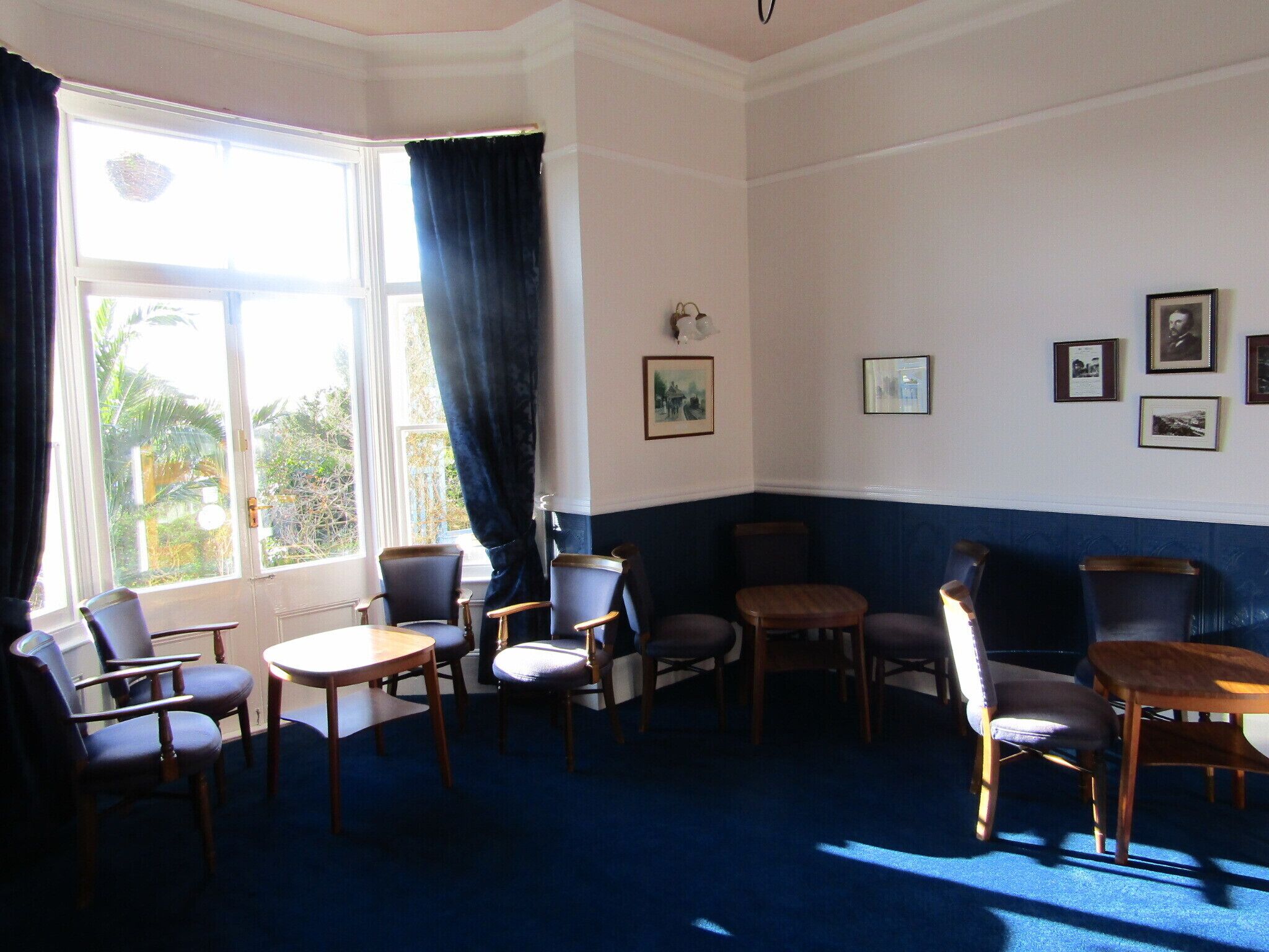 A room with a bay window, several chairs, a table, and framed pictures on the wall.