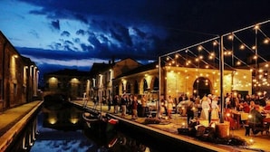 A riverside restaurant with outdoor seating, a canal, and a brick building in the background. - Michelangelo Holiday & Family Resort (Comacchio)