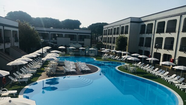 A swimming pool with lounge chairs and umbrellas, surrounded by buildings. - Michelangelo Holiday & Family Resort (Comacchio)