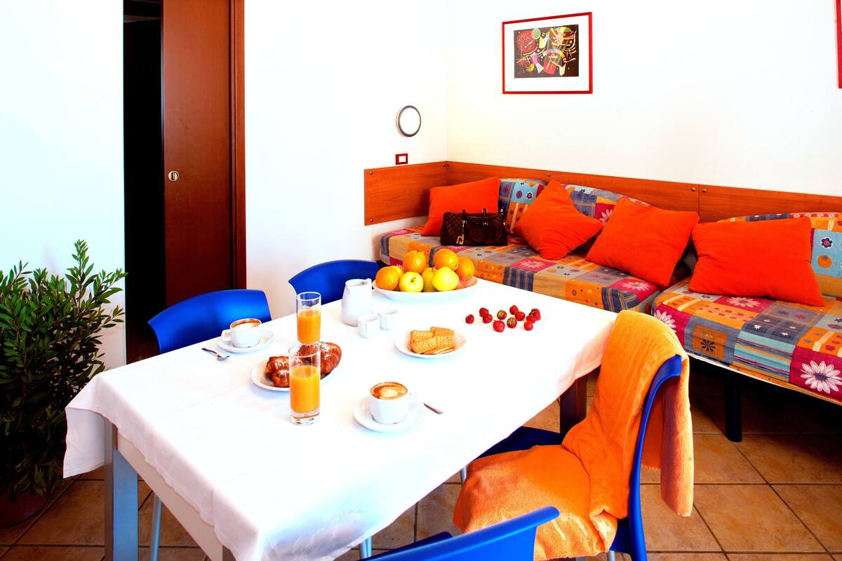 A dining area with a table set for breakfast, including plates of fruit, pastries, and beverages. There is a sofa with orange cushions in the background.