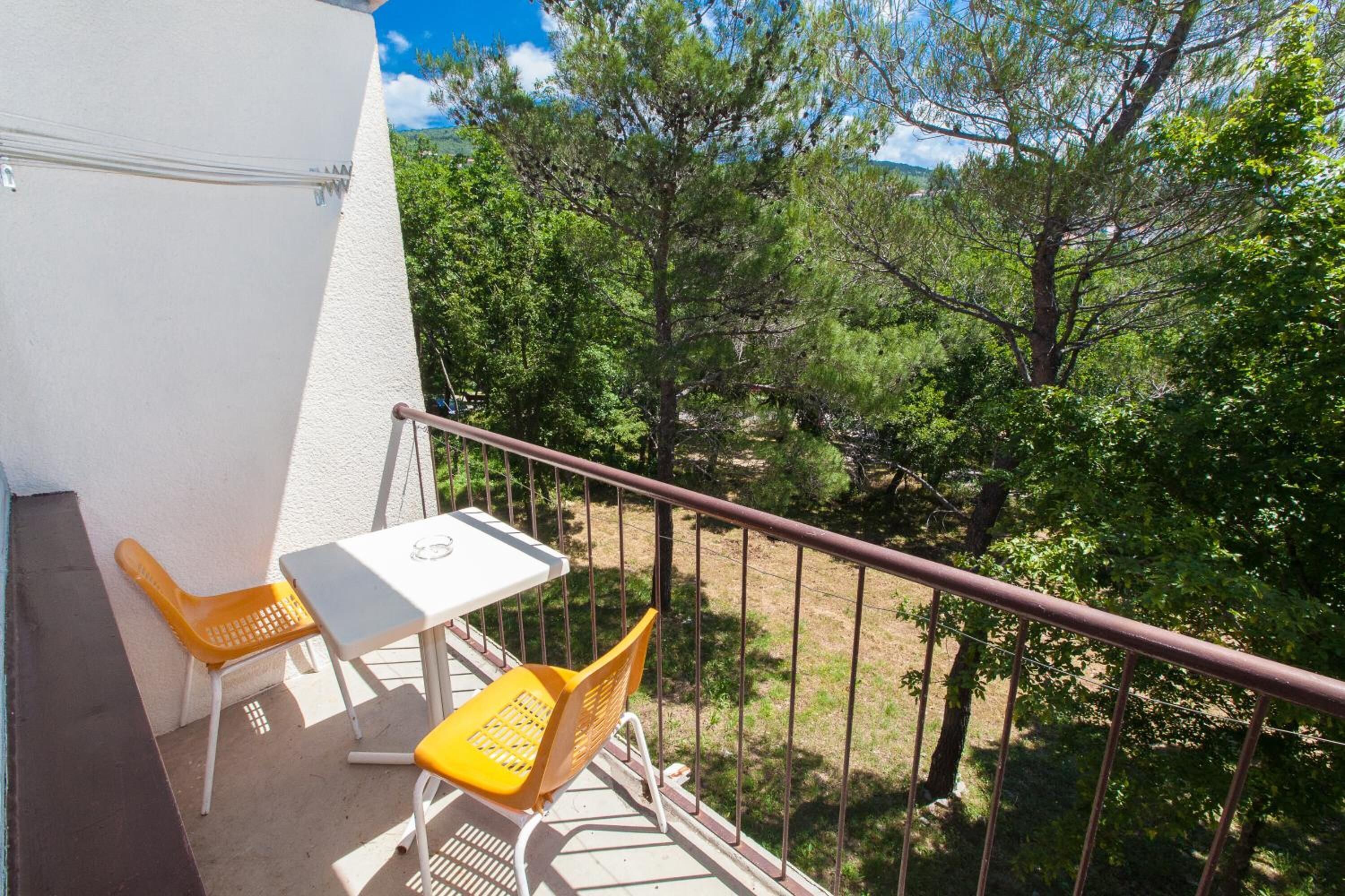 Varanda com uma mesa branca e duas cadeiras amarelas, com vista para uma área arborizada.