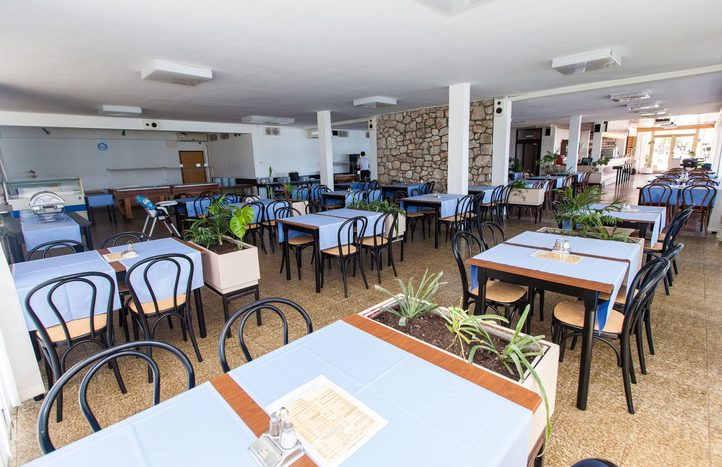 A spacious indoor dining area with multiple tables and chairs, a kitchen in the background, and a stone wall.