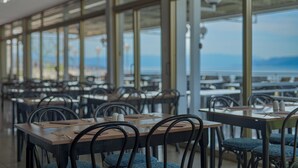 Un restaurante con mesas y sillas dispuestas en filas, ventanas amplias y vista al mar.