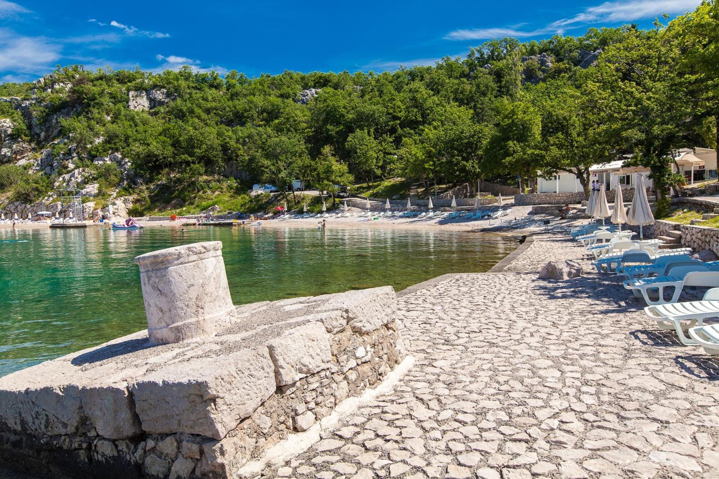 A coastal area with a stone-paved walkway, beach umbrellas, and lounge chairs along the water's edge.
