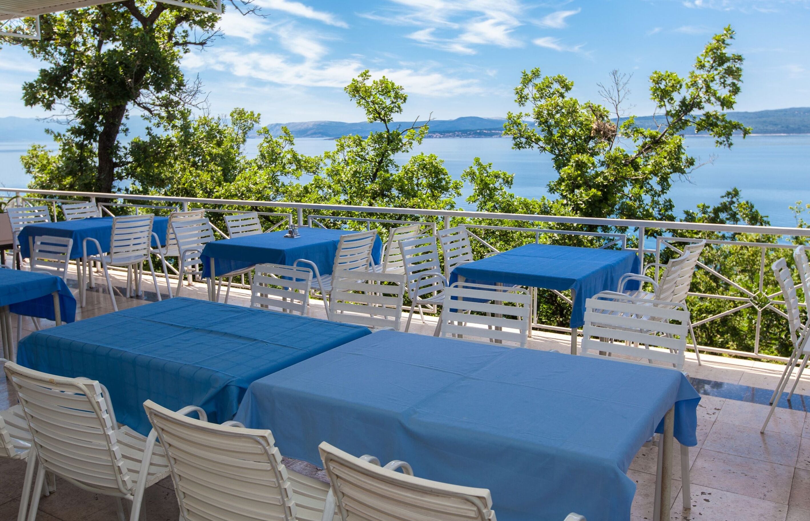 A terrace with tables and chairs, overlooking a body of water and hills.