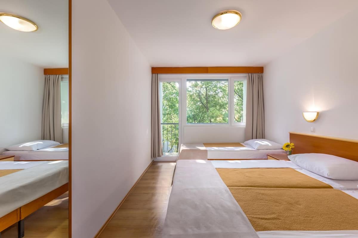 Habitación de hotel con dos camas, un ventanal con vistas a árboles y una mesita de noche con lámpara.
