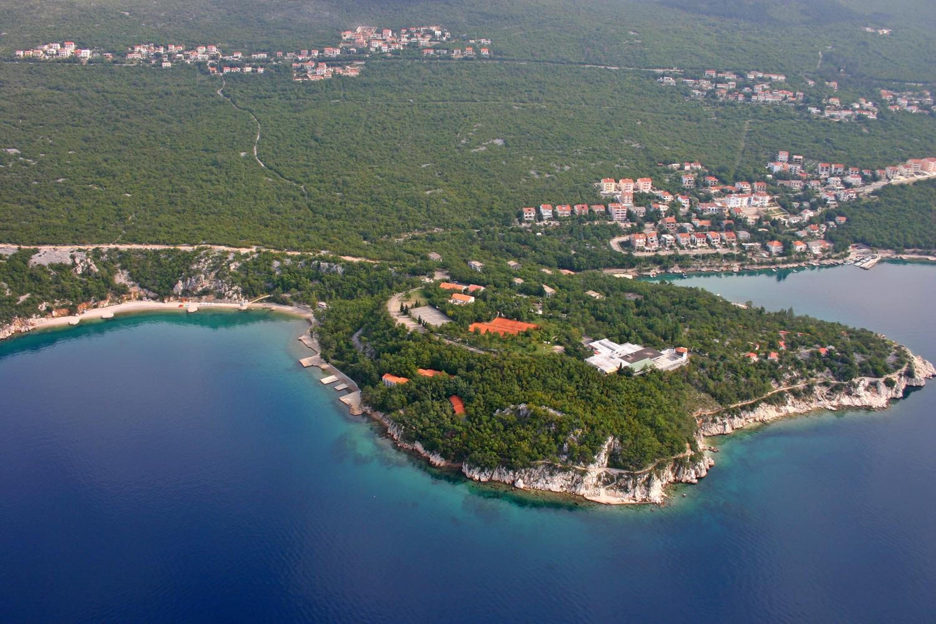 An aerial view of a coastal town with a central cluster of buildings surrounded by lush greenery and a curved coastline.