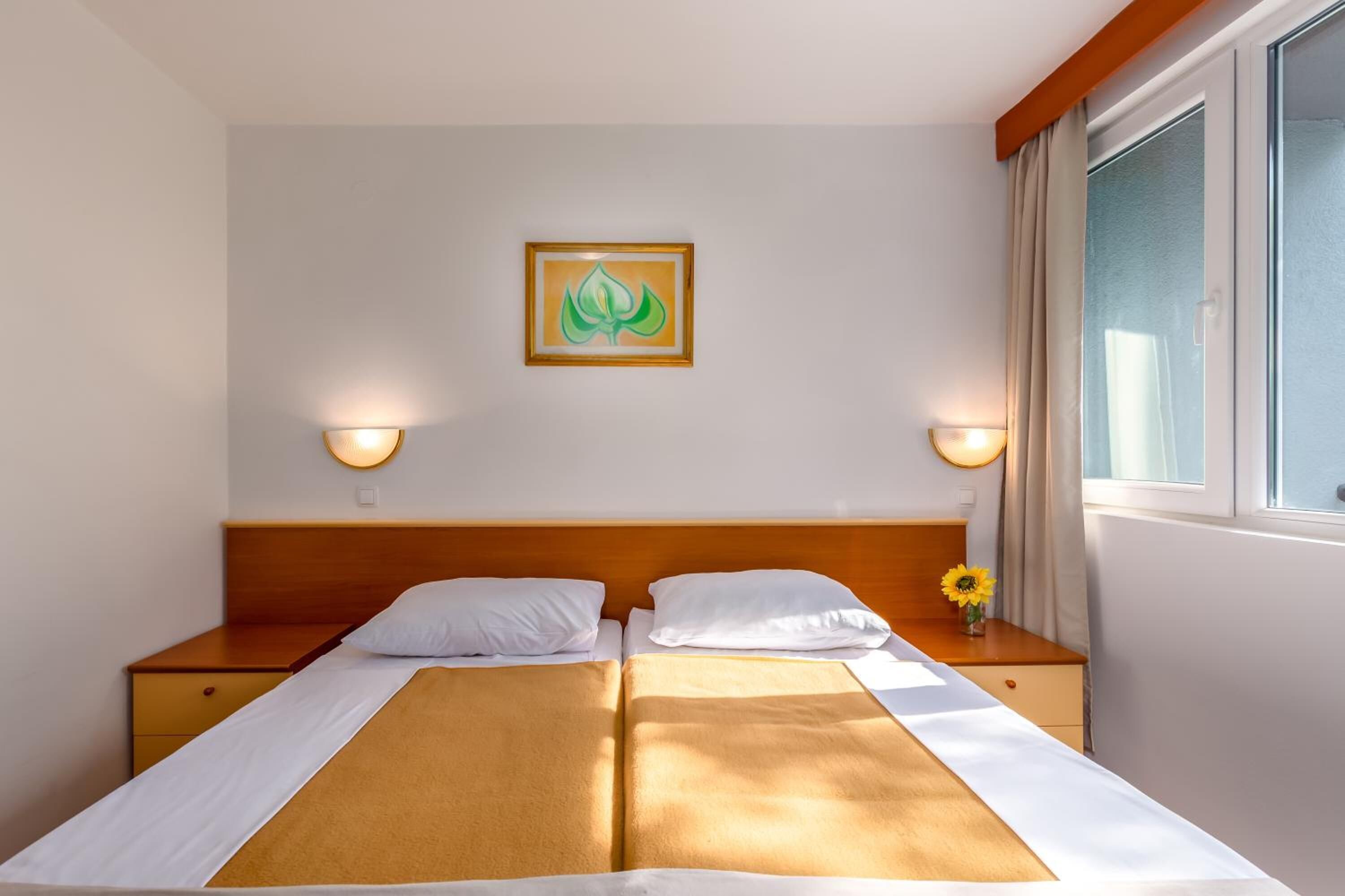 Cama doble con cabecera de madera, dos mesitas de noche, lámpara de pared y ventana con cortinas.