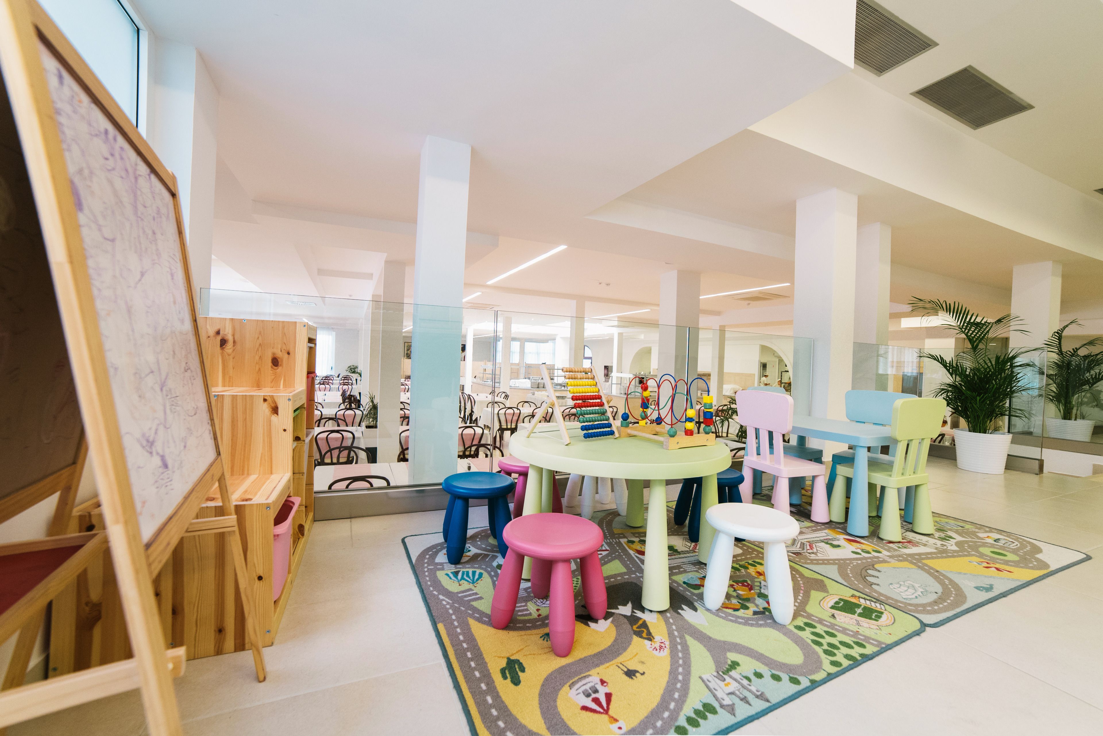Un espacio interior bien iluminado con una mesa redonda rodeada de taburetes y sillas coloridos, un caballete de madera y juguetes infantiles.