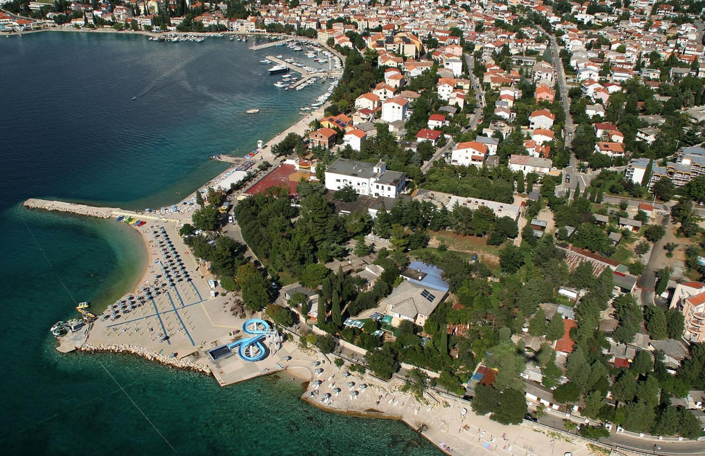 An aerial view of a coastal town with a marina, residential buildings, and a beach area with umbrellas and a water slide.