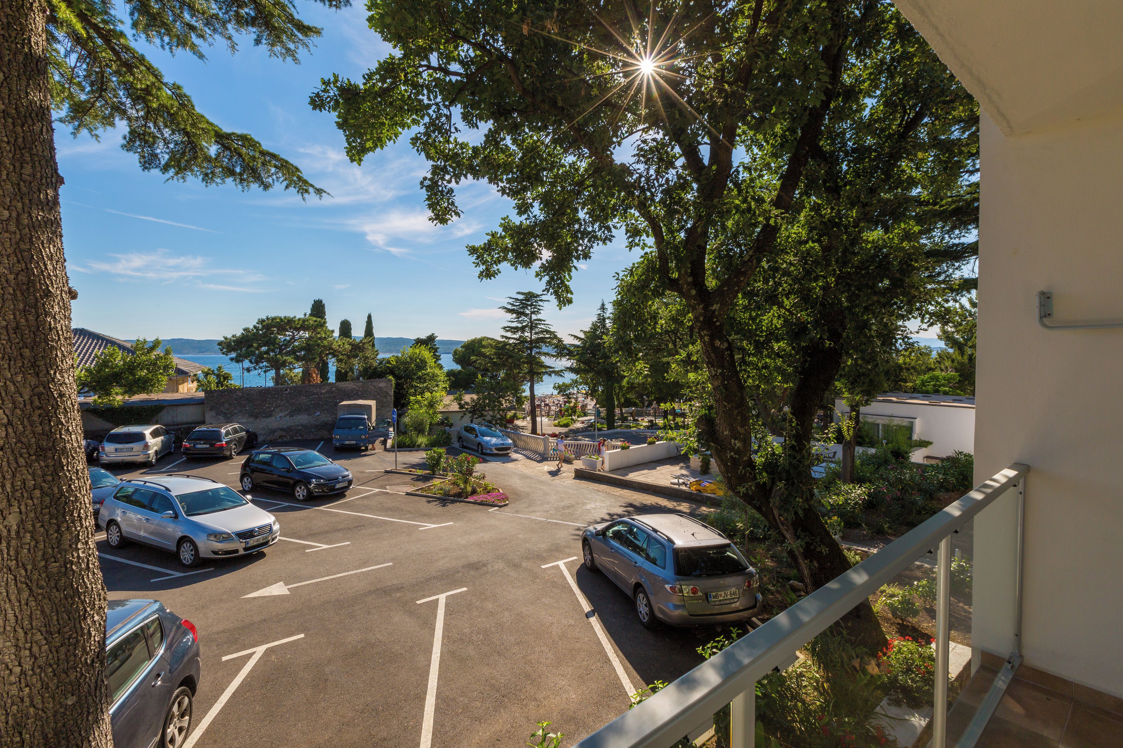 A parking area with multiple cars, a view of the sea, and a building with a balcony overlooking the parking lot.