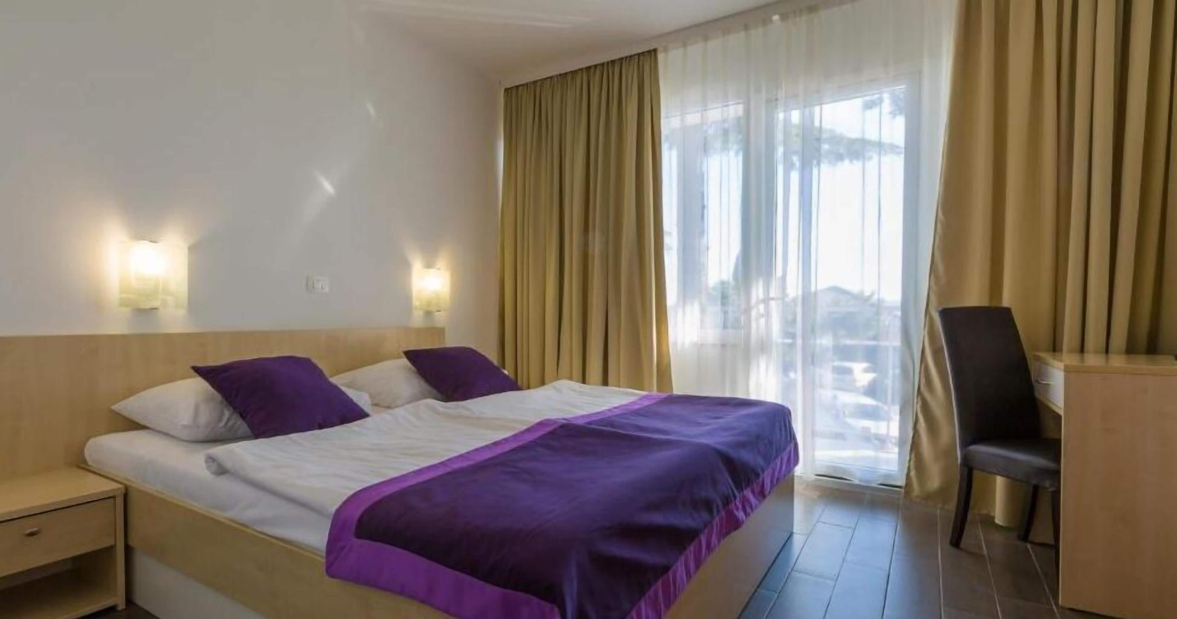 Hotellihuone, jossa on sänky, kaksi yöpöytää, työpöytä, tuoli ja suuri ikkuna, jossa on verhot.