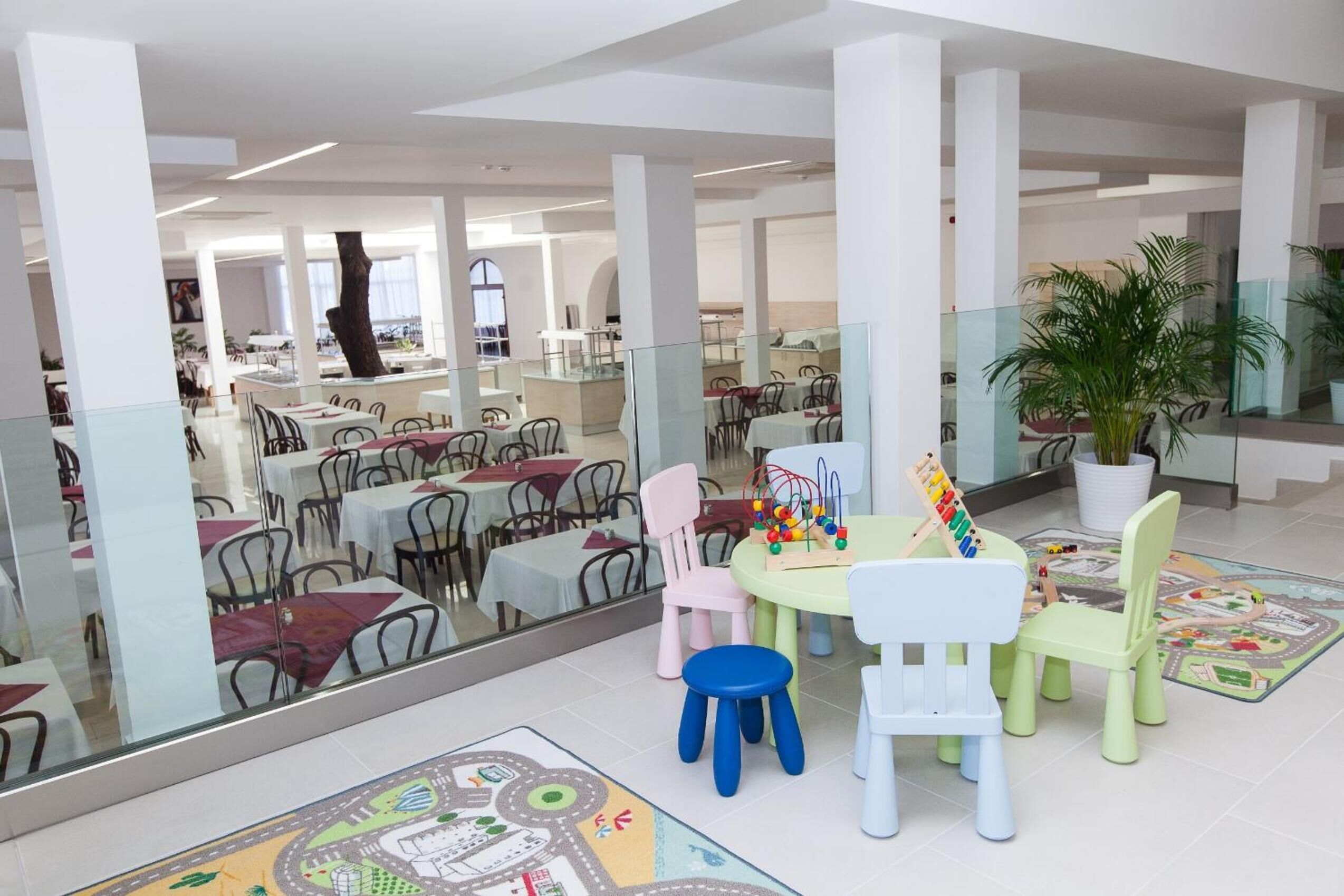 A spacious indoor area with multiple tables and chairs, a children's play area with toys, and a large potted plant.