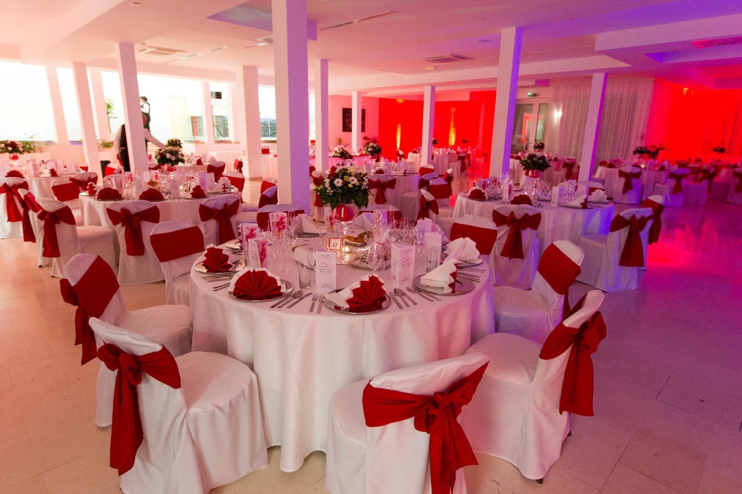 A banquet hall with round tables set for a formal event, white tablecloths, and red sashes on chairs.