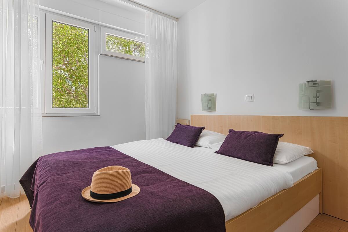 A bedroom with a bed, a hat on the bed, a window with curtains, and a wall-mounted light fixture.