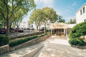 Zona de recepción de un hotel con un camino empedrado que lleva a la entrada, rodeada de árboles y coches aparcados.