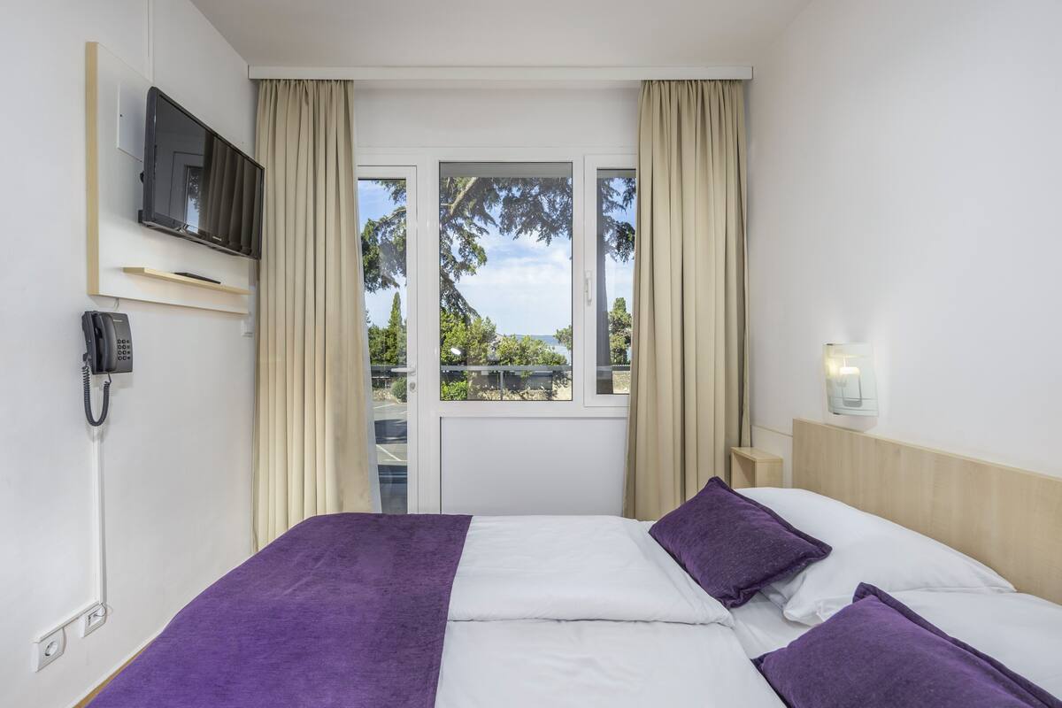 A hotel room with a bed, a TV mounted on the wall, a phone, and a window with curtains.