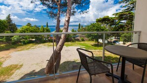 A balcony with a table and chairs, overlooking a sandy area with trees and a view of the sea. - Hotel Slaven (Crikvenica)