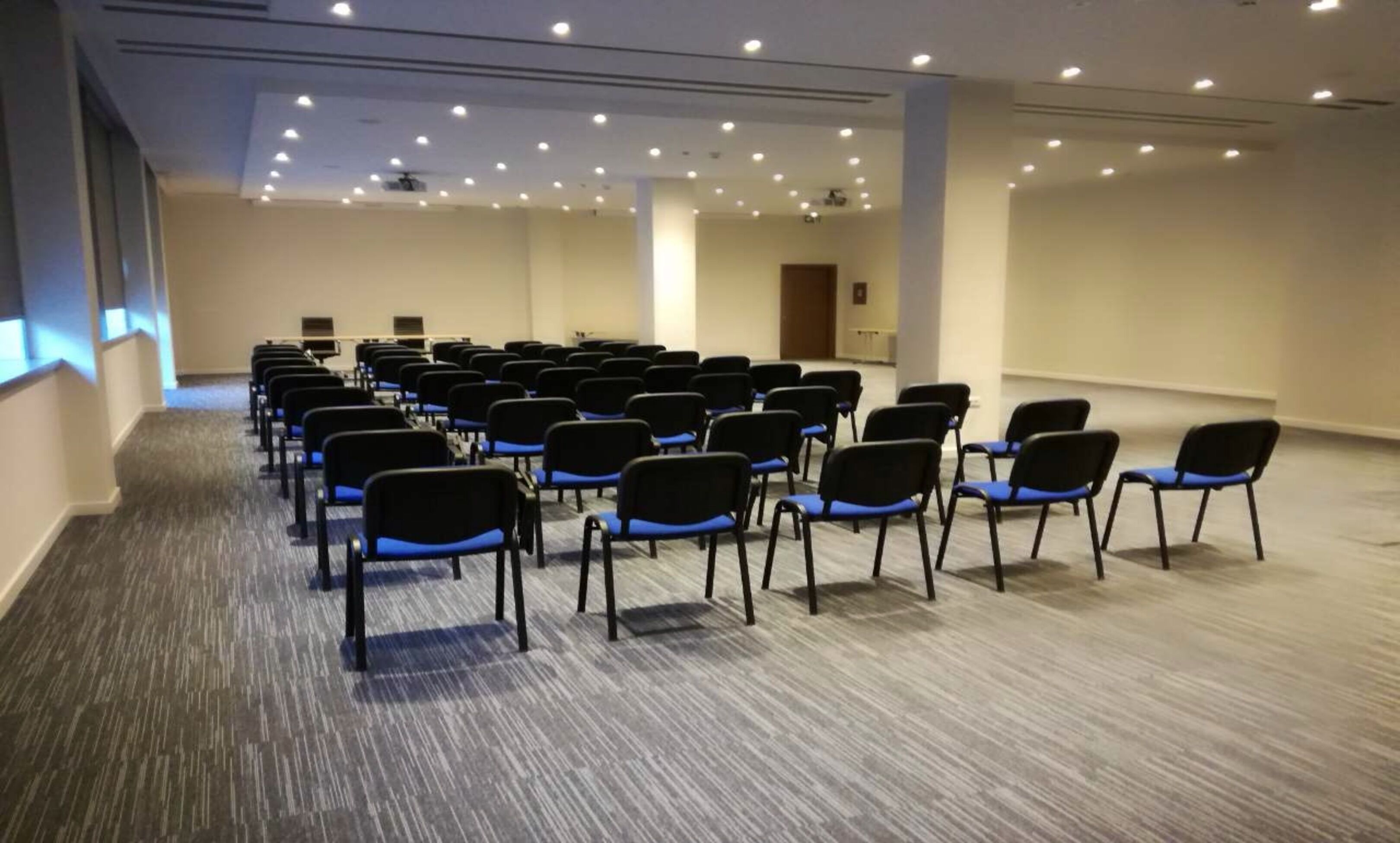 A conference room with rows of black chairs with blue cushions, carpeted floor, and recessed ceiling lights.