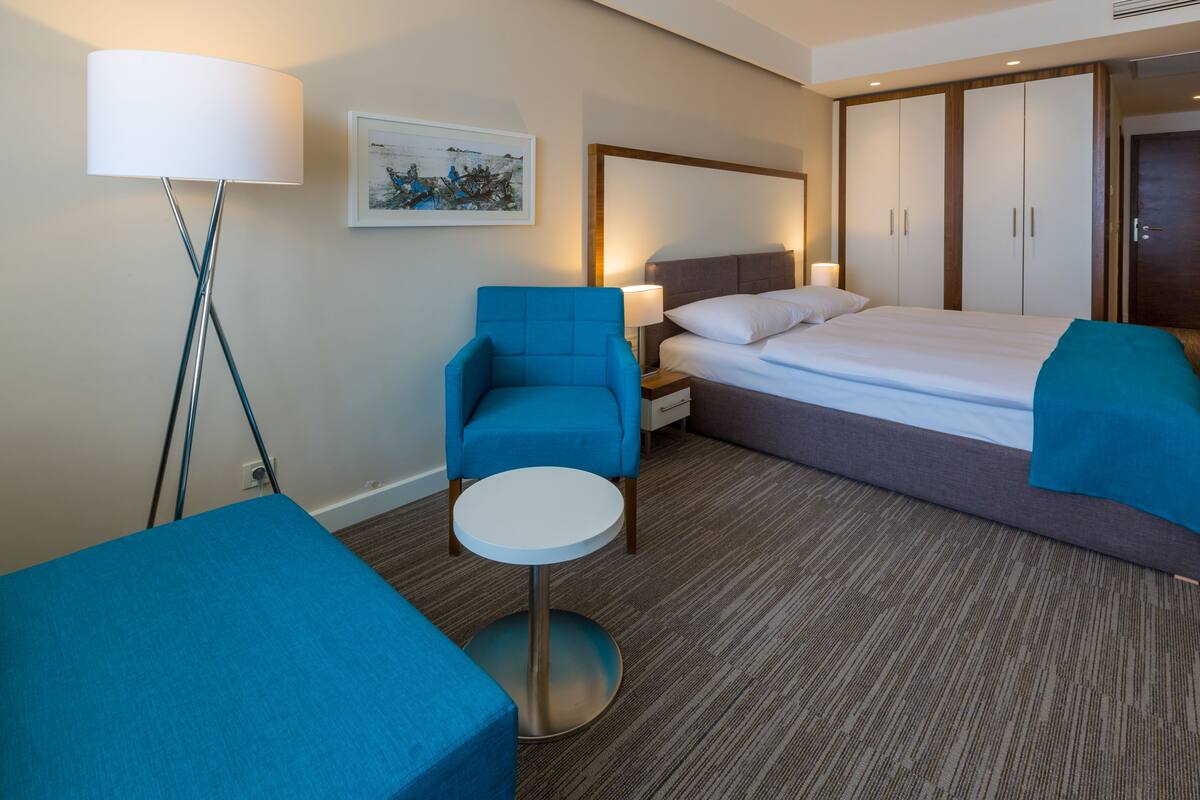 A hotel room with a bed, a blue armchair, a small round table, a floor lamp, and a painting on the wall.