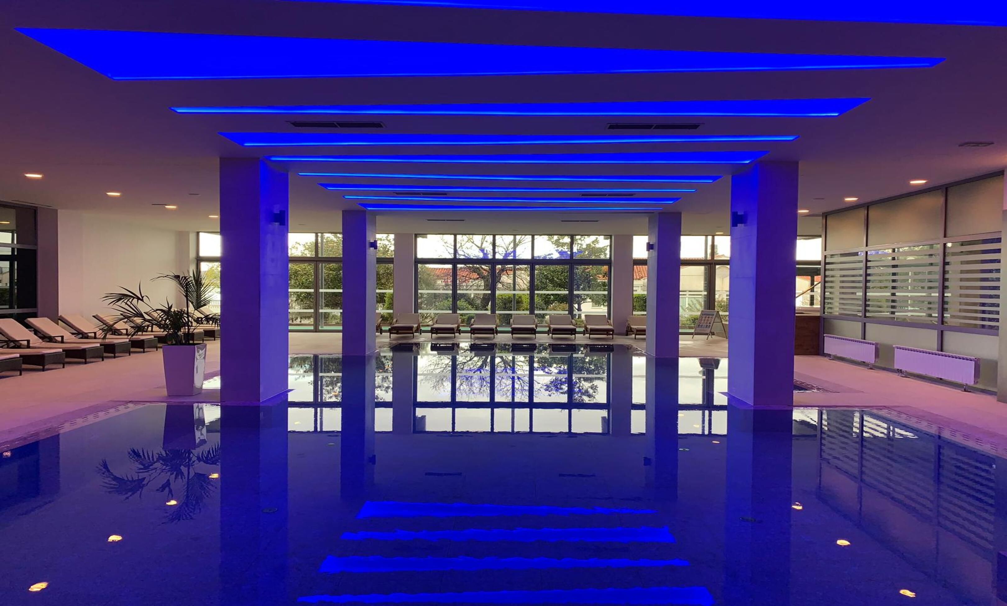 A modern indoor pool area with a clear view of the pool, lounge chairs, and a glass wall with a view of the outside.