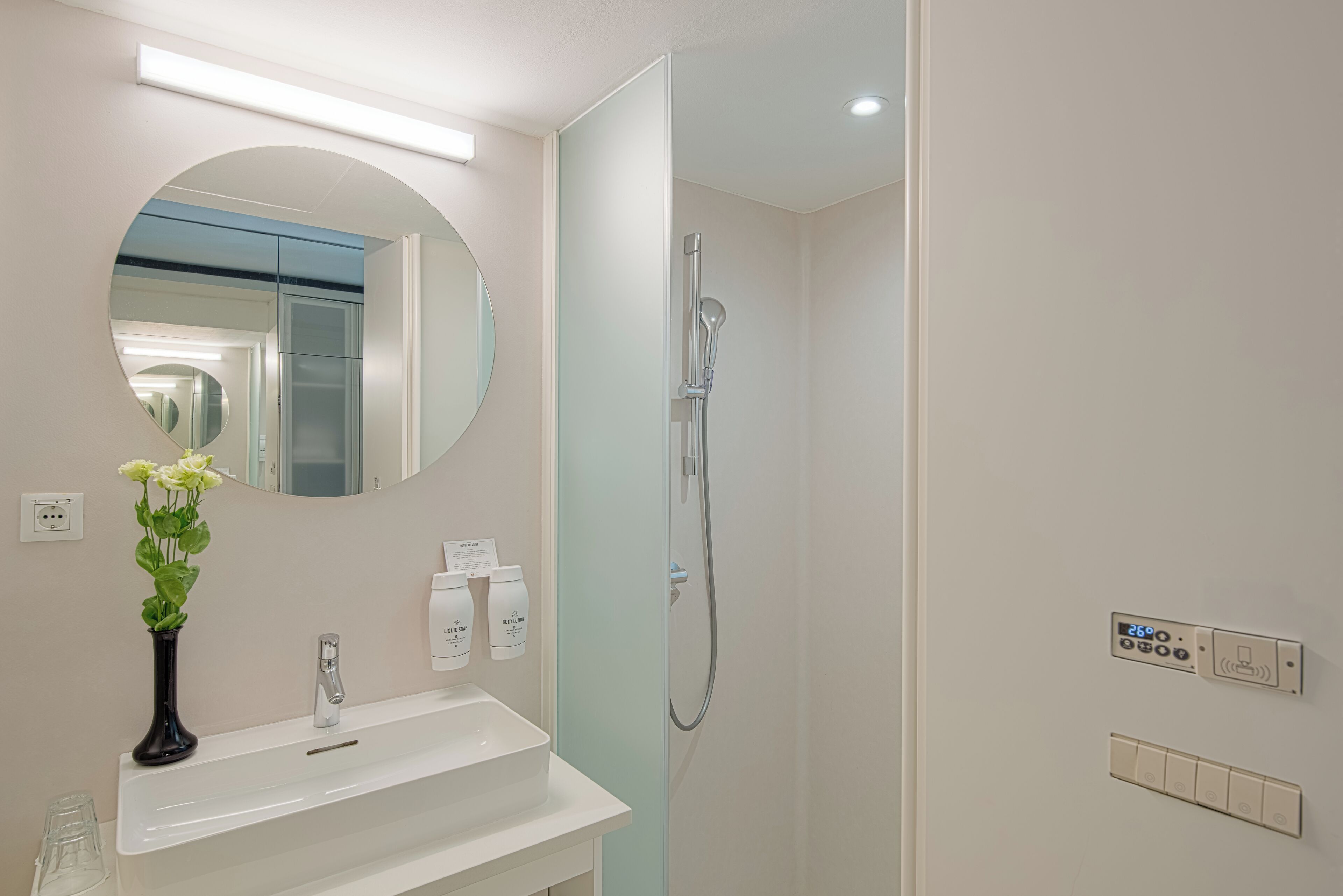 A modern bathroom with a white sink, a mirror, a shower, and a wall-mounted control panel.