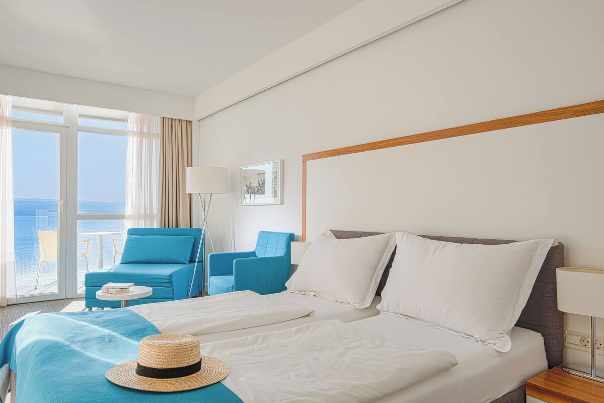 A hotel room with a bed, two blue chairs, a small table, a lamp, and a view of the sea.