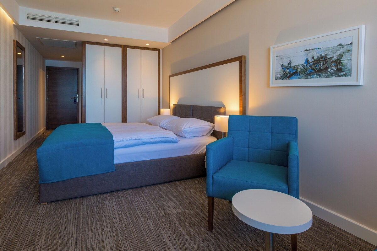 A hotel room with a bed, a chair, a small table, and a painting on the wall.