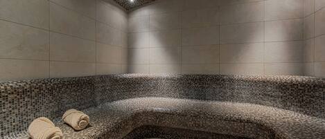 A modern spa room with a mosaic-tiled bench, two rolled towels, and a recessed ceiling light.