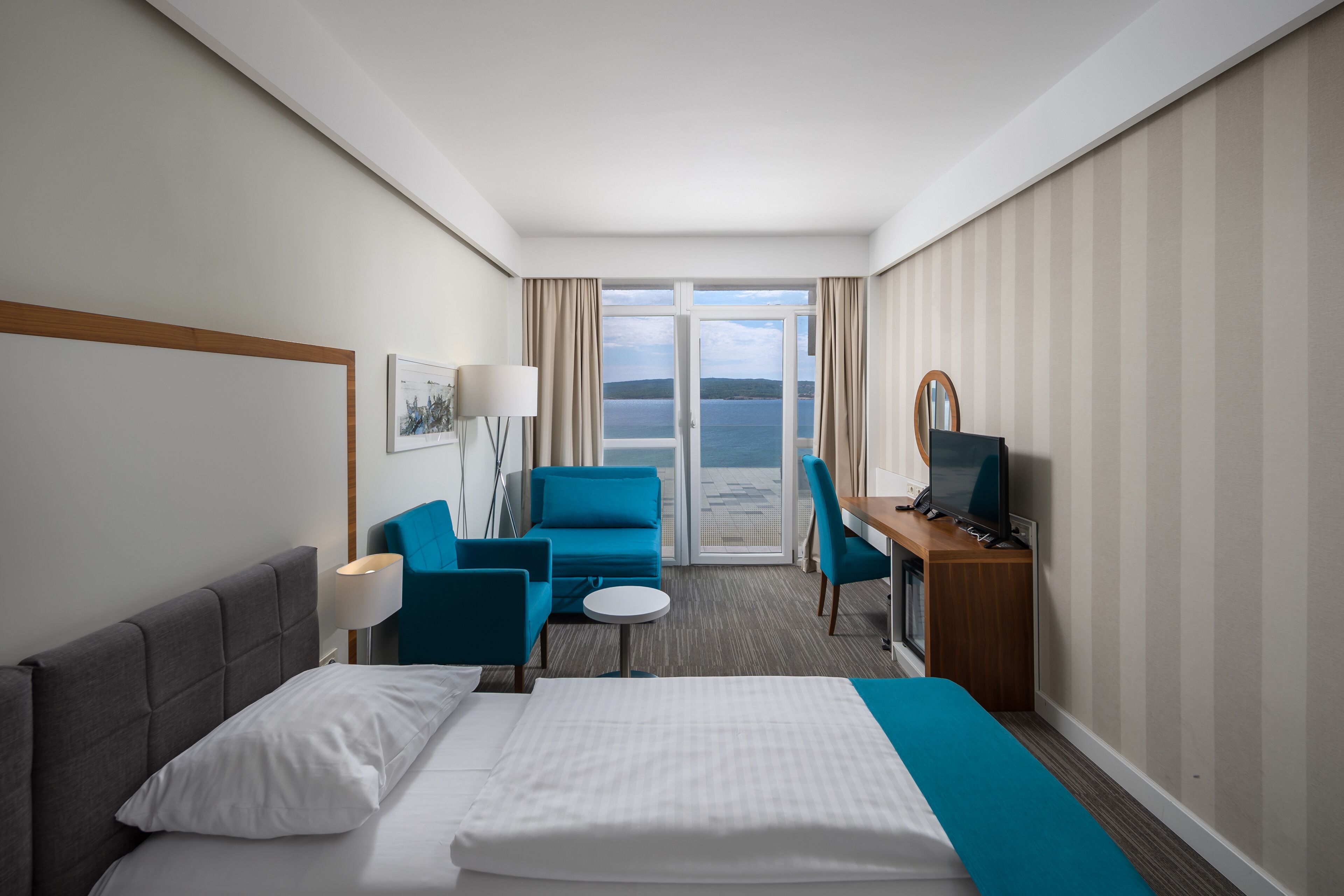 A hotel room with a bed, a desk with a computer, a chair, a small table, and a view of the sea through a large window.