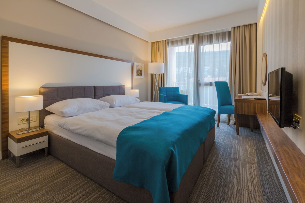 A hotel room with a large bed, a desk with a chair, a television, and a window with curtains.