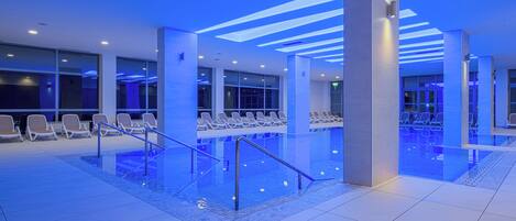 A modern indoor pool area with clear glass railings, lounge chairs, and blue ambient lighting.
